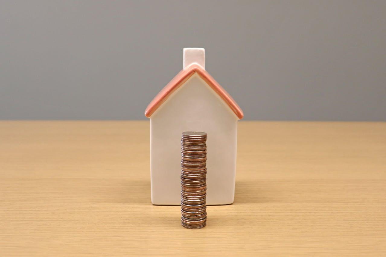Small white house with a red roof on a wooden table, with a pile of coins in front of it.