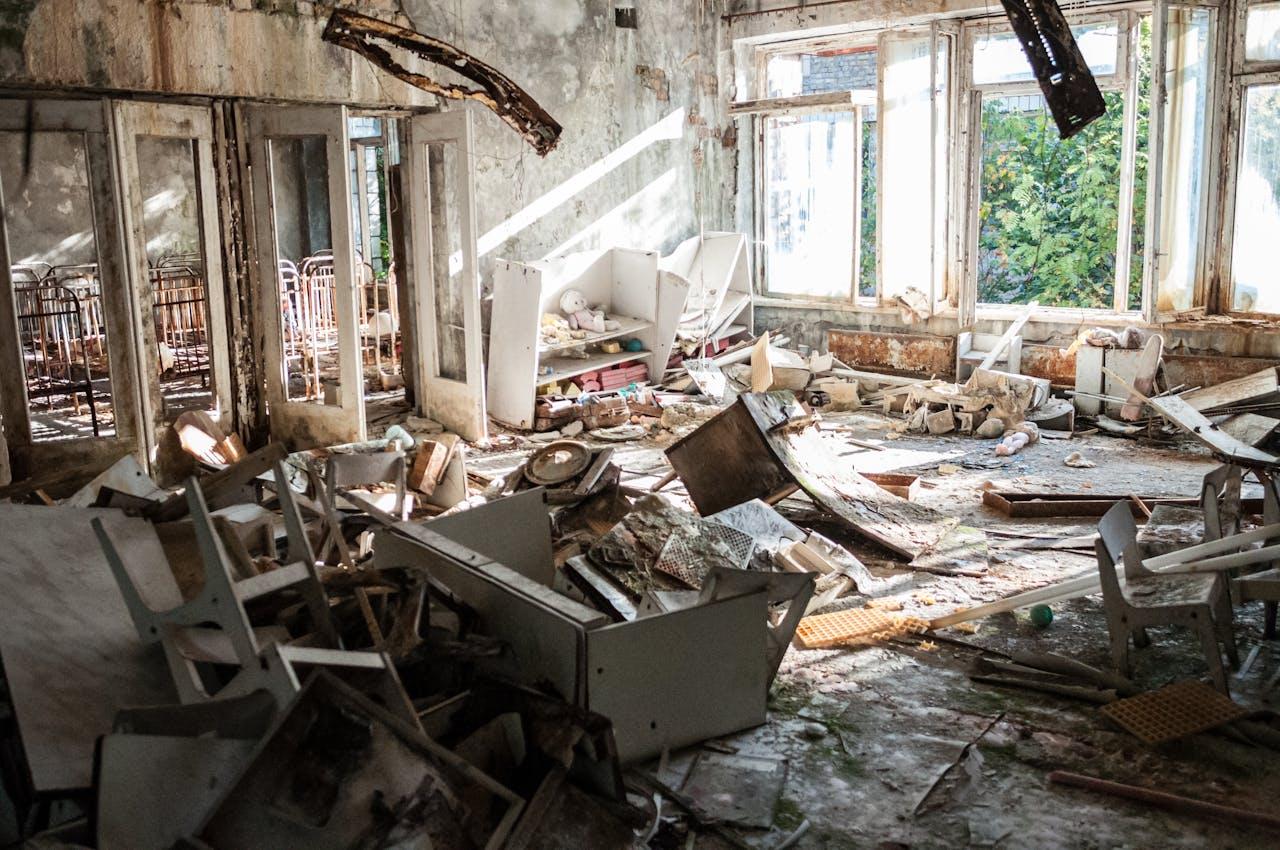 Abandoned room with rubble, broken furniture, and rusted bed frames, illuminated by sunlight from broken windows.