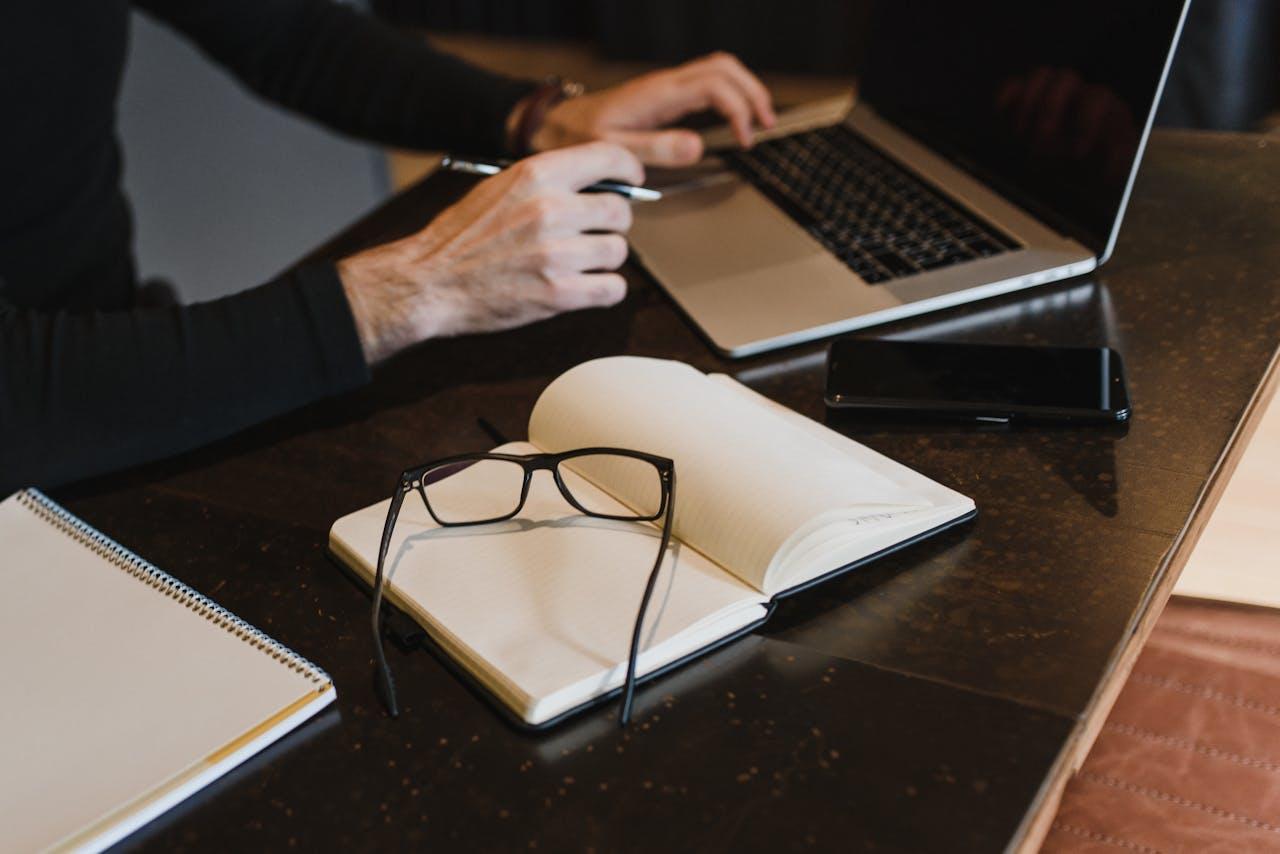 Ein Schreibtisch mit einem offenen Laptop, einem Smartphone, einem aufgeschlagenen Notizbuch mit einer Brille darauf und einem weiteren Notizblock. Eine Person in schwarzer Kleidung arbeitet am Laptop und haelt einen Stift in der Hand.