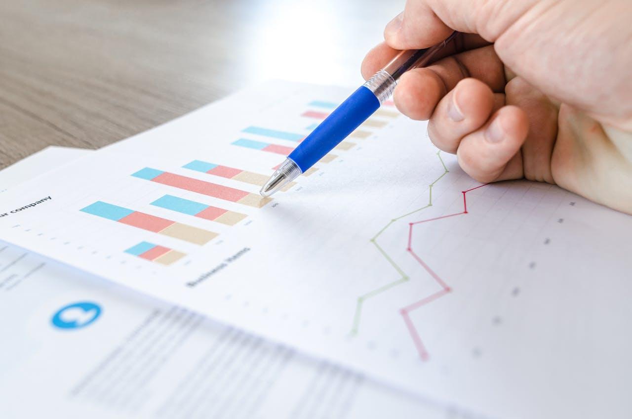 A hand holds a ballpoint pen with a blue grip and points to a printed sheet of paper with bar charts in blue, red and beige and a line chart in red and green. The document is on a wooden table.