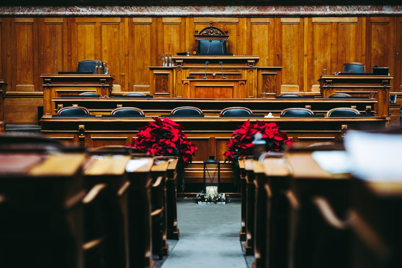 Holzvertäfelter parlamentarischer Sitzungssaal mit einer erhöhten Richter- oder Sprecherbank in der Mitte, umgeben von leeren schwarzen Stühlen. Weihnachtssterne (rote Pflanzen) und eine Laterne schmücken den Raum.