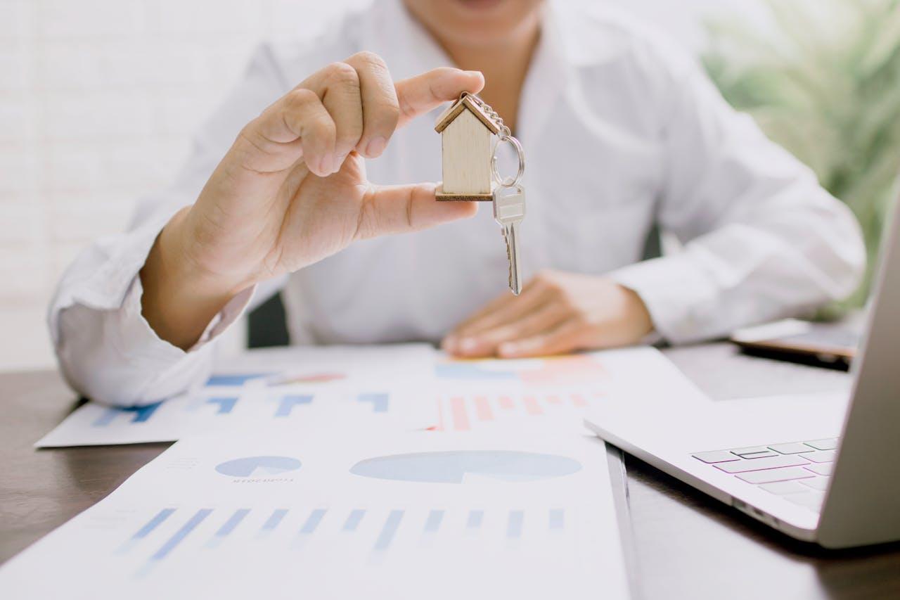 Eine Person in einem weissen Hemd hält einen Schlüsselbund mit einem kleinen Holzhaus-Anhänger. Auf dem Schreibtisch liegen Finanzdiagramme, ein Laptop und weitere Unterlagen, was auf Immobiliengeschäfte oder Investitionen hinweist.